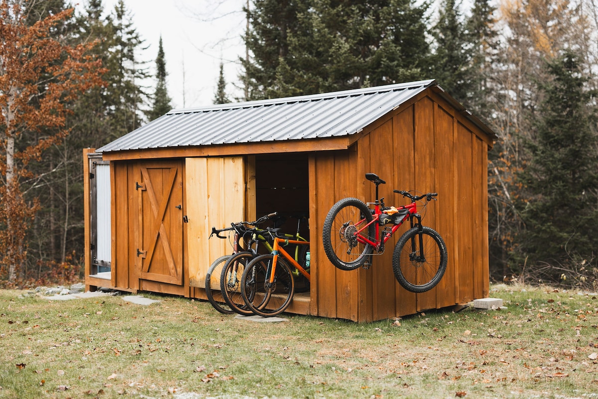 A wooden shed with a metal roof is situated on grassy ground, surrounded by trees. The structure features two sliding doors and is equipped with bike storage. Several bicycles are mounted on the wall and leaned against the exterior, highlighting convenient access for outdoor enthusiasts.
