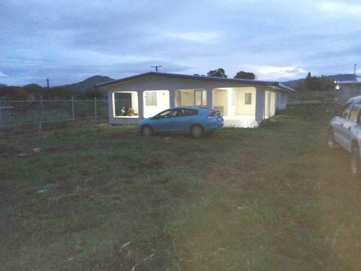 An exterior view of a single-story home is shown during twilight, featuring a blue car parked in the foreground. The house has multiple windows that reflect the evening light, while a grassy area surrounds the structure.