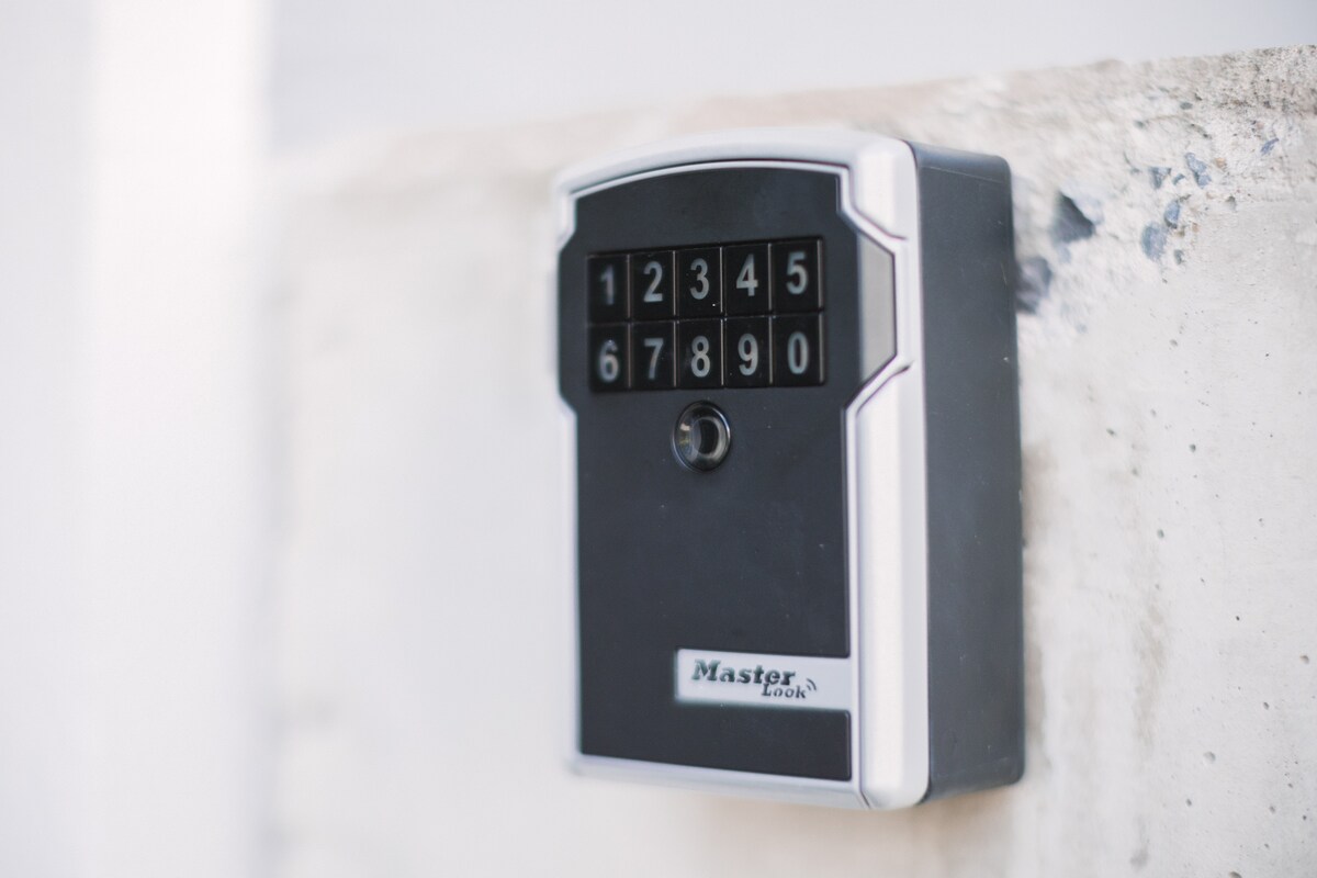 A wall-mounted key lockbox is displayed, featuring a numerical keypad and a key slot. The lockbox is installed on a textured concrete surface, providing secure access to the apartment for guests. Its design enables contactless entry for convenience.