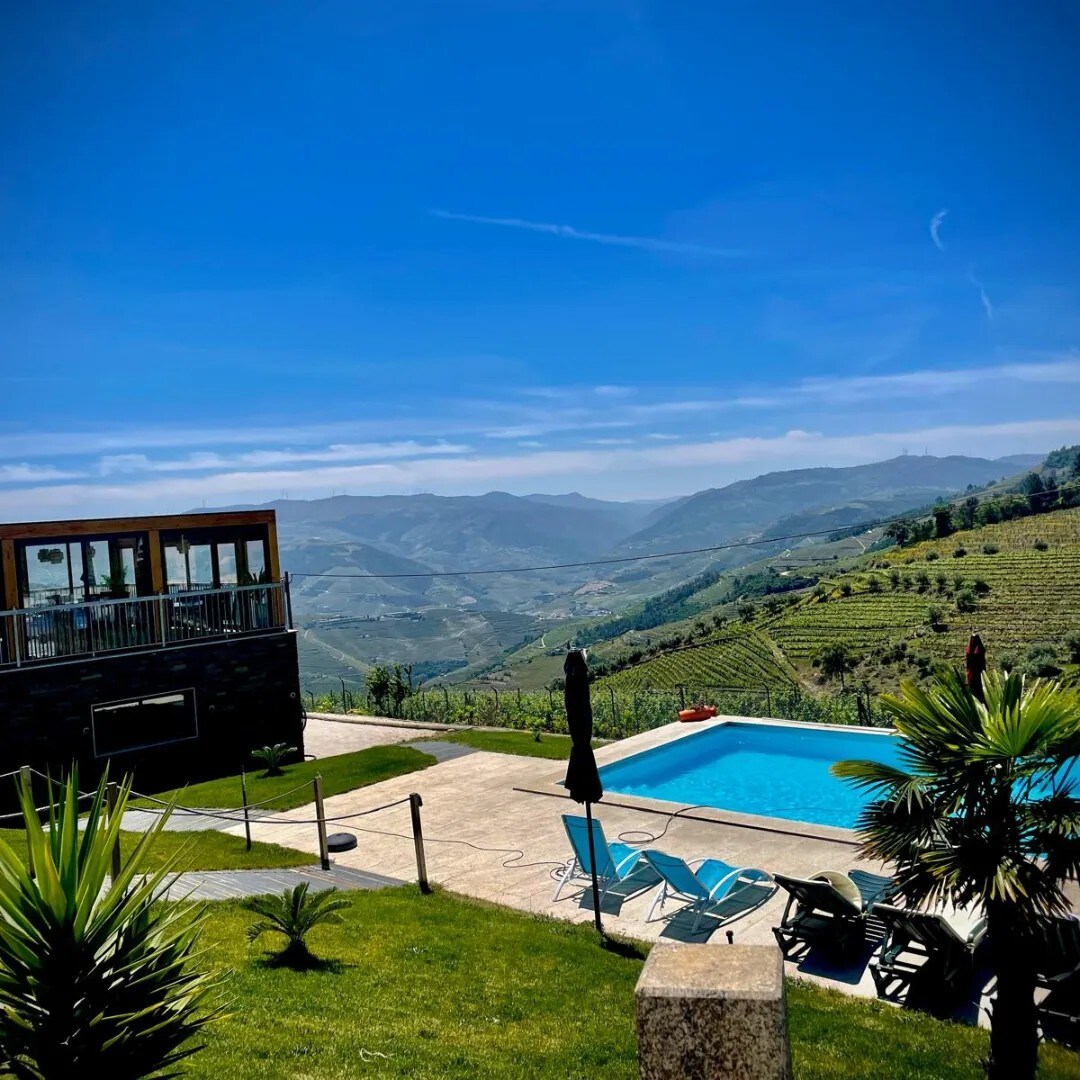 A spacious outdoor area features a refreshing pool surrounded by sun loungers and greenery. A wooden structure with large windows overlooks scenic valleys, while distant hills provide a picturesque backdrop under a clear blue sky.