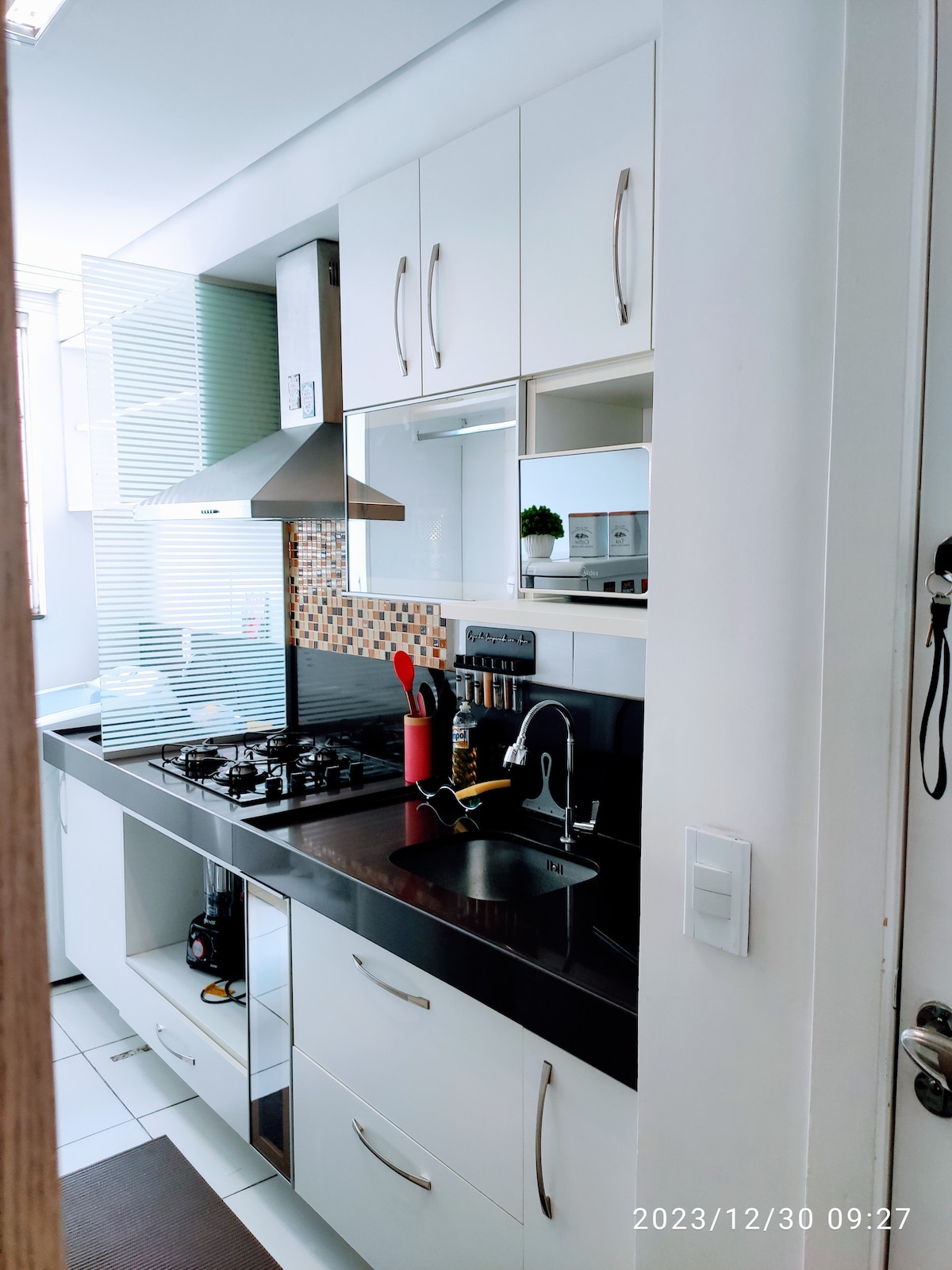 A modern kitchen features an efficient layout with sleek white cabinetry and contrasting dark countertops. A built-in gas stove and sleek range hood are visible. Organized storage spaces and decorative tiles add a contemporary touch, while natural light brightens the cooking area through a nearby window.