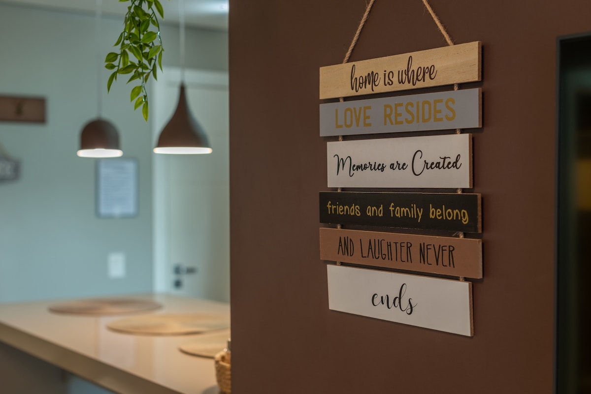 A decorative wall hanging features a series of wooden slats with text expressing themes of love, memories, friendship, and laughter. The slats are in various earthy tones, providing a warm touch to the interior space. Soft lighting is visible in the background.