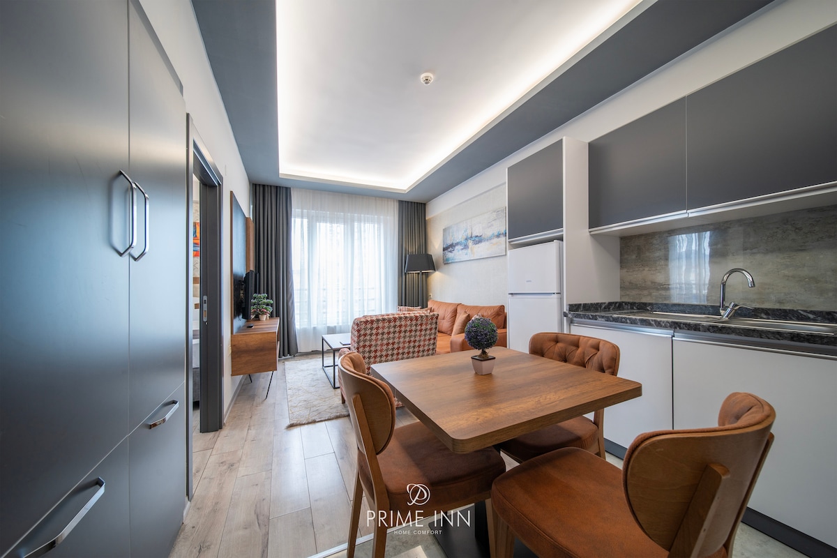 A modern living area is highlighted, featuring a comfortable seating arrangement with a sofa. A dining table with four chairs is positioned near the kitchen, which includes a refrigerator and cooking area. Natural light filters through large windows, creating a bright, inviting space.
