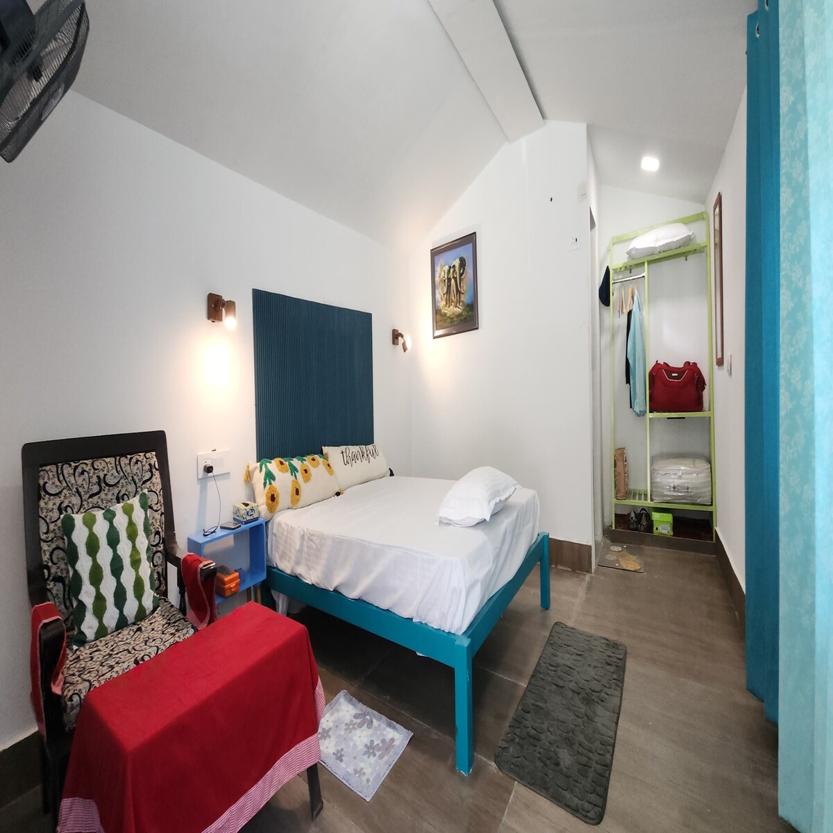 A bright, airy room features a spacious bed with crisp linens. A colorful armchair and a small red table are present, along with a wardrobe that offers storage. Soft lighting complements the decor, and a rug adds warmth to the space.