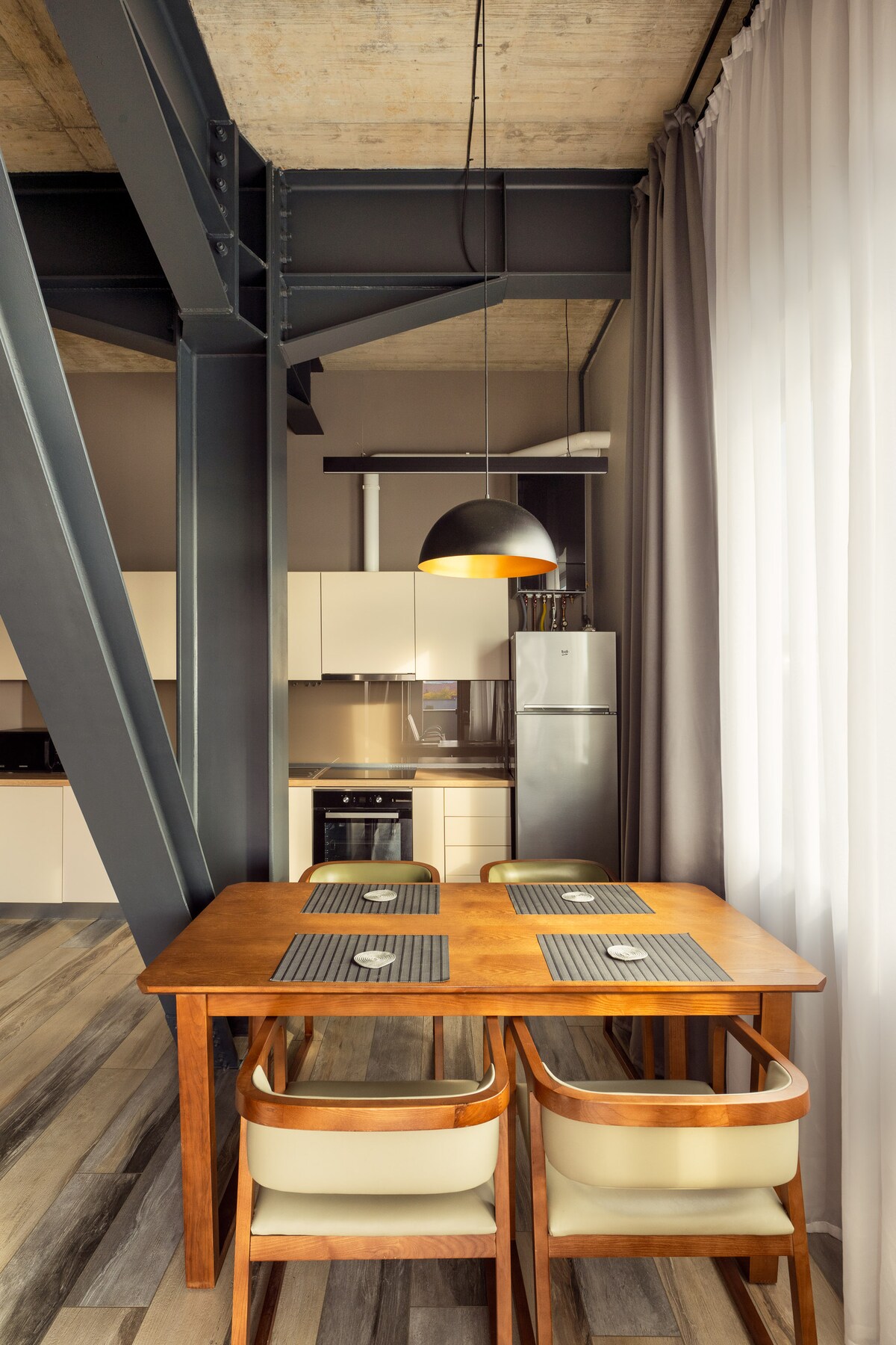 A dining area features a wooden table accompanied by two chairs, positioned near the kitchen. A pendant light hangs above, providing illumination. The backdrop includes modern cabinetry and a stainless steel refrigerator, complemented by large windows dressed with light gray curtains.