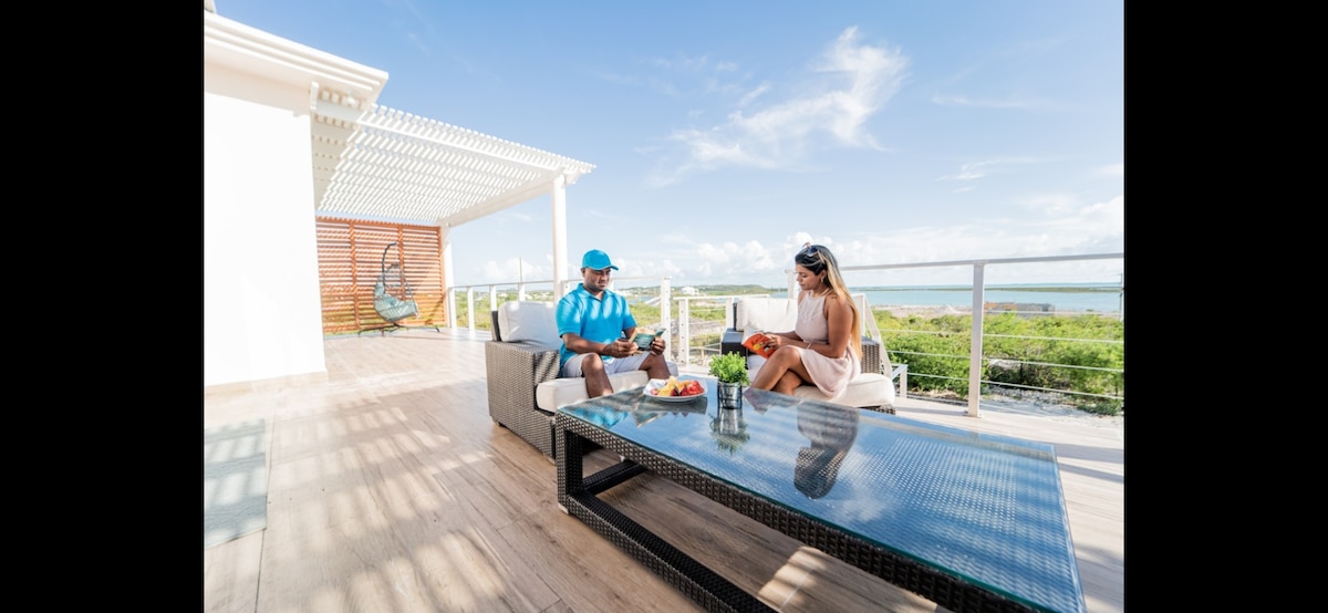 A sheltered outdoor space features a modern seating arrangement, consisting of a sofa and a coffee table. A couple is seated, enjoying refreshments with a scenic backdrop of lush greenery and a clear blue sky. The setting offers a relaxed atmosphere for social interaction.
