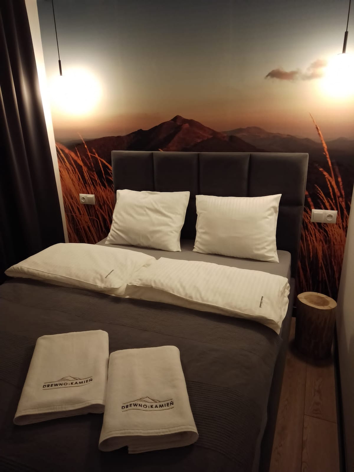 A cozy sleeping area features a comfortable bed with two plush pillows. The backdrop showcases a serene mountain landscape, while soft lighting hangs above. Neatly arranged towels are placed at the foot of the bed, adding to the inviting setting.