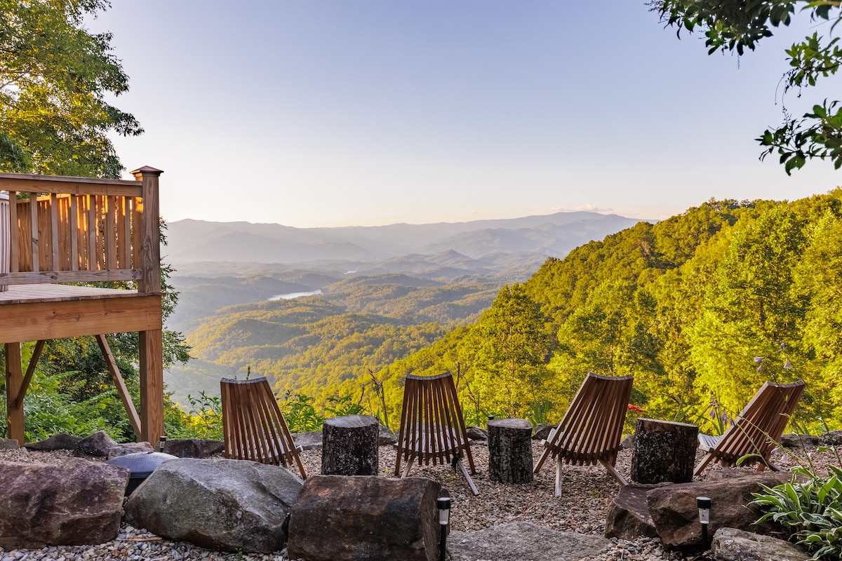 A wooden deck overlooks a stunning mountain view, with multiple chairs arranged in a semi-circle. The landscape features rolling hills covered in lush green foliage, extending into the distance. Sunlight casts a gentle glow over the serene environment, creating a peaceful retreat setting.