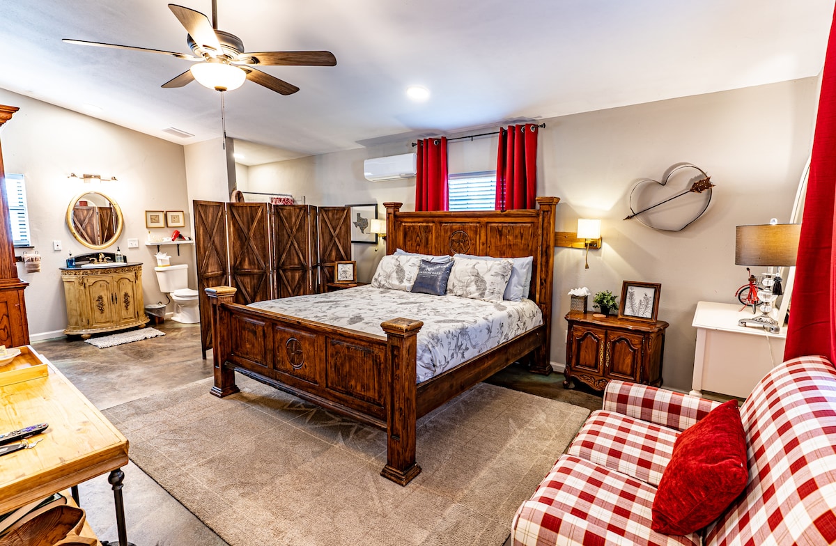 The spacious room features a large wooden bed with a patterned comforter. Natural light filters through red curtains, and a cozy armchair with a checkered pattern is positioned nearby. A wooden desk and an elegant screen provide additional privacy, complementing the warm tones of the decor.