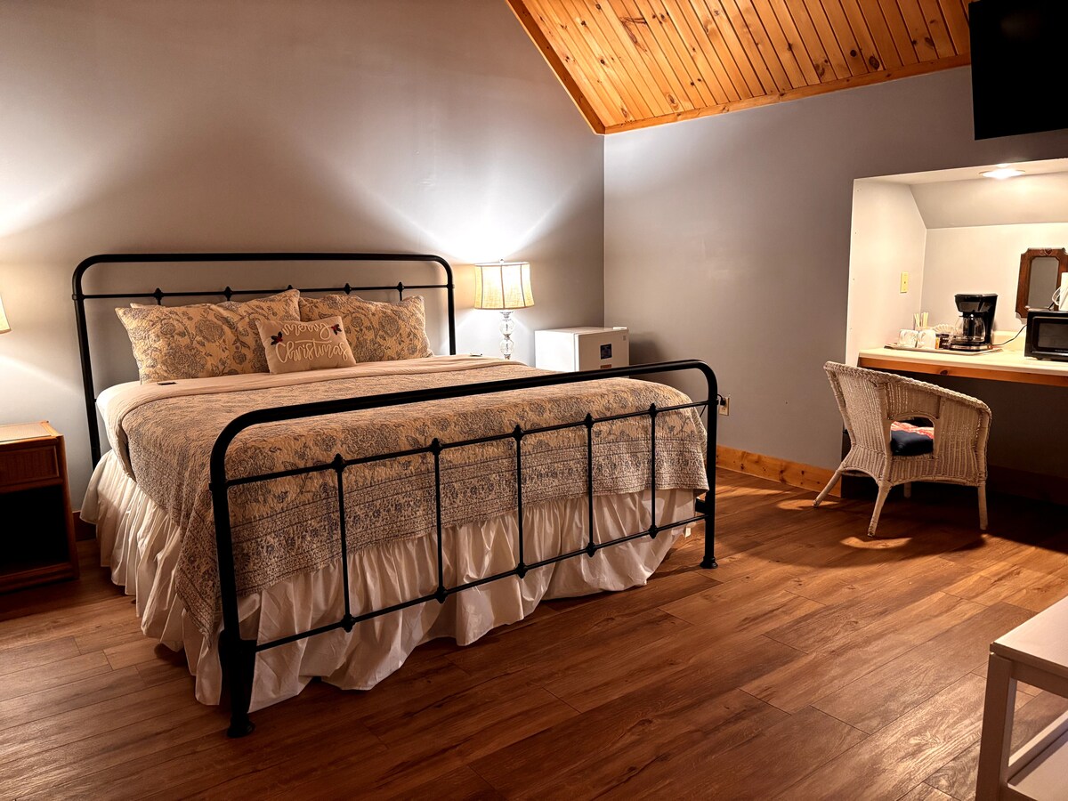 A well-appointed room features a king-sized bed with decorative pillows and a light-colored bedspread. A small desk with a chair is positioned near the window, and a coffee maker is also present. Soft bedside lamps provide ambient lighting against wooden flooring.