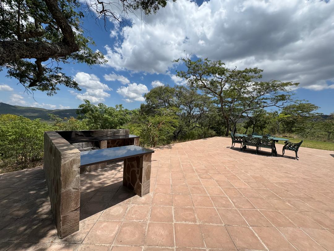 An outdoor area surfaces with rustic tiles, featuring a stone barbeque grill and a dining table with metal chairs. Surrounding greenery and open skies create a serene environment, inviting guests to enjoy nature.