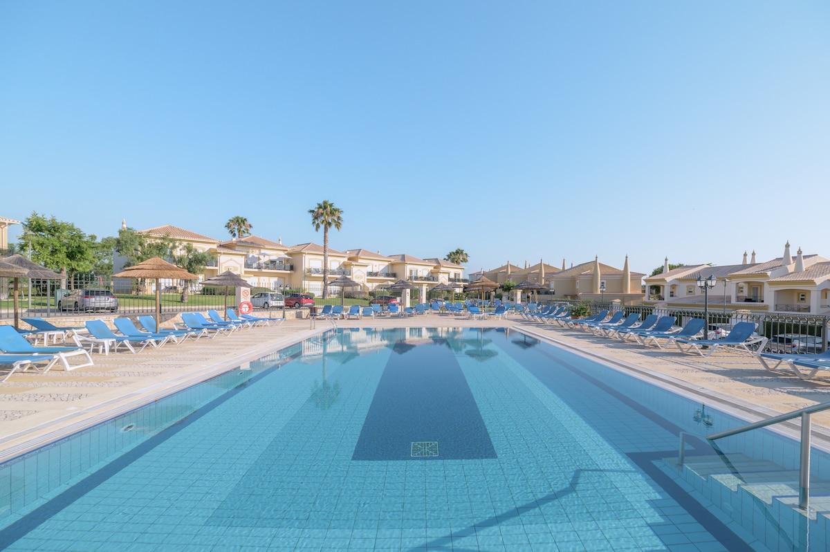A large outdoor pool features crystal-clear water surrounded by a spacious sunbed area. Lounge chairs arranged neatly around the pool are complemented by palm trees in the background, creating a serene environment. The resort's buildings are visible in the distance under a clear blue sky.