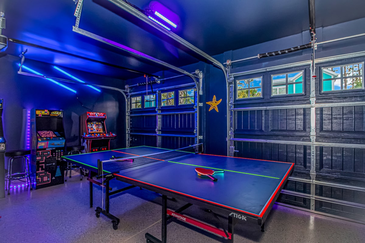 The brightly lit garage game room features a ping pong table with vibrant blue and red accents. To one side, several arcade games are available, complemented by neon lighting and a starfish decoration on the wall, creating an engaging atmosphere for entertainment.