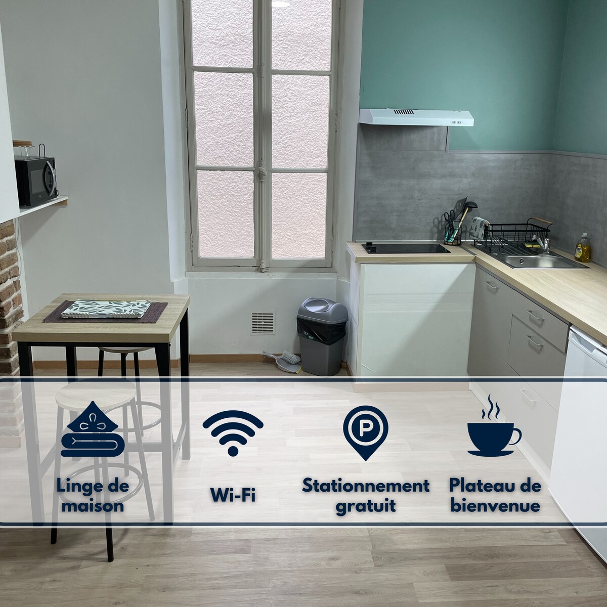 The kitchen area features a well-equipped layout with modern appliances and a light color scheme. A small dining table is positioned next to the window, allowing natural light. Essential items such as a garbage bin and countertop items are also visible.