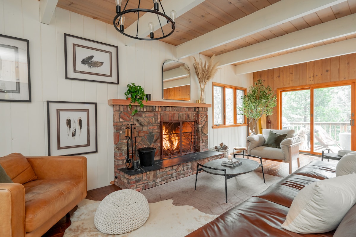A welcoming living room features a stone fireplace with a warm glow, surrounded by leather and fabric seating. Large windows provide natural light and views of greenery outside. Decorative art pieces add character, while a round pouf and coffee table complete the inviting space.