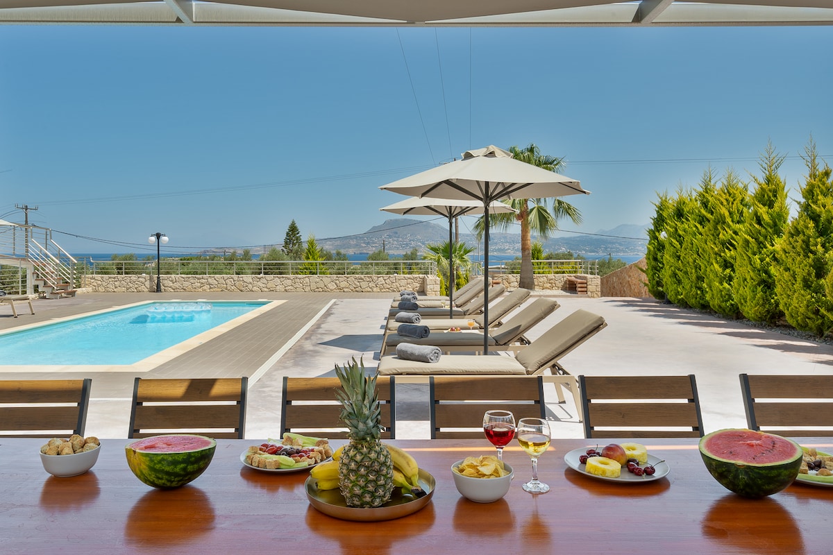 A large dining table is set outdoors, adorned with an assortment of fresh fruits and refreshments. In the background, a private pool surrounded by lounge chairs offers a serene setting, complemented by shaded umbrellas and lush greenery on either side.