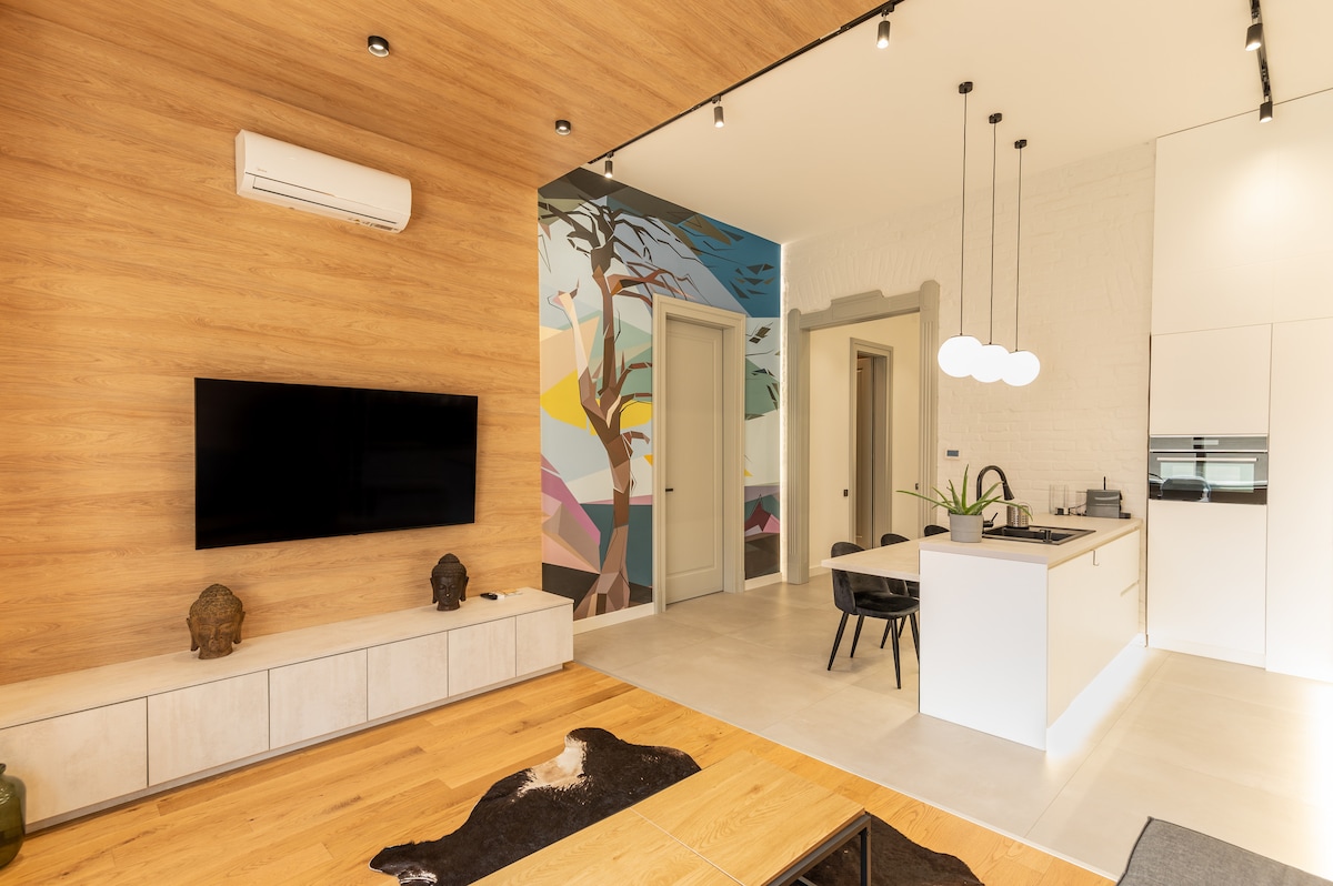 A modern living area is highlighted by a large wall-mounted television against a warm wooden panel. A vibrant mural depicting nature adds color to one wall. The kitchen features sleek cabinetry and a central island with seating, while contemporary lighting fixtures enhance the space.