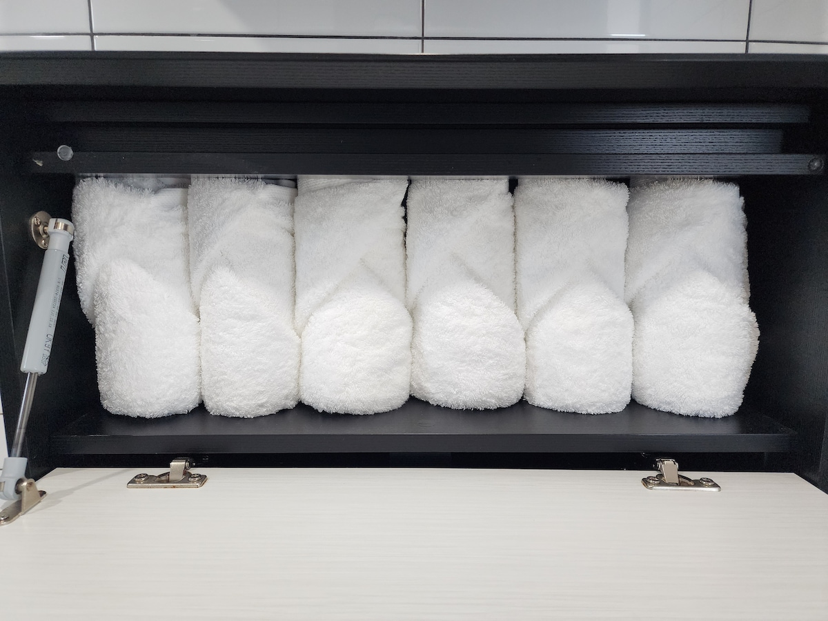A neatly arranged shelf displays six rolled white towels, each wrapped with care. The towels are placed on a dark shelf, contrasting with the light-colored surface below. This organized storage provides easy access for guests.