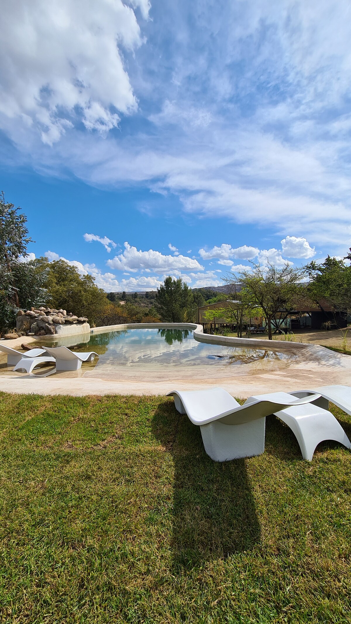 A spacious outdoor area features a unique pool surrounded by a lush, grassy landscape. White lounge chairs are positioned nearby, inviting relaxation under a bright blue sky filled with fluffy clouds. Trees and a distant structure can be seen, enhancing the natural setting.