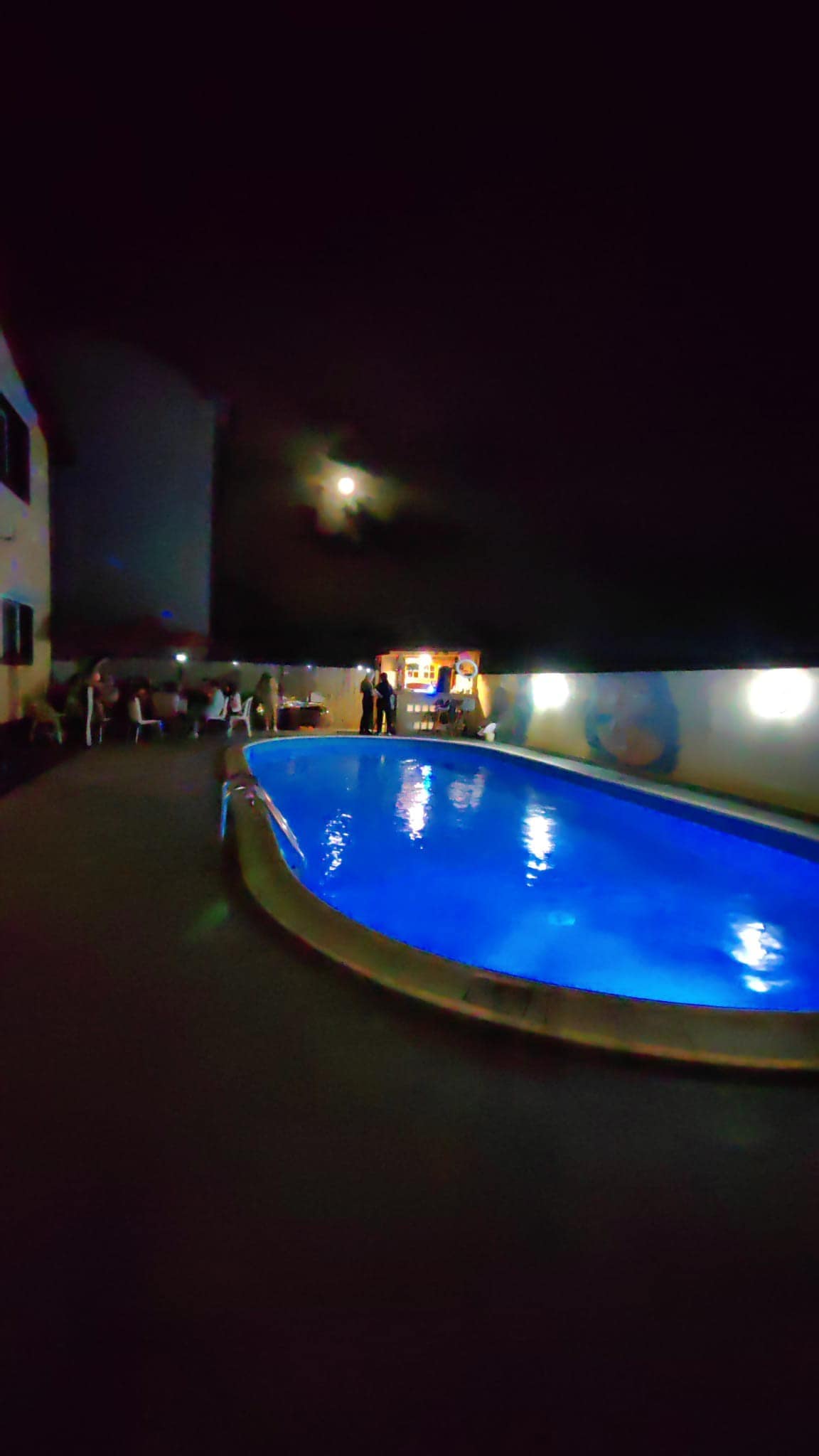 The outdoor pool, illuminated by soft blue lights, reflects the night sky and surrounding area. Guests are seen enjoying the space, with the pool bar in the background offering a welcoming atmosphere. The pool's curved edge complements the building's architecture.