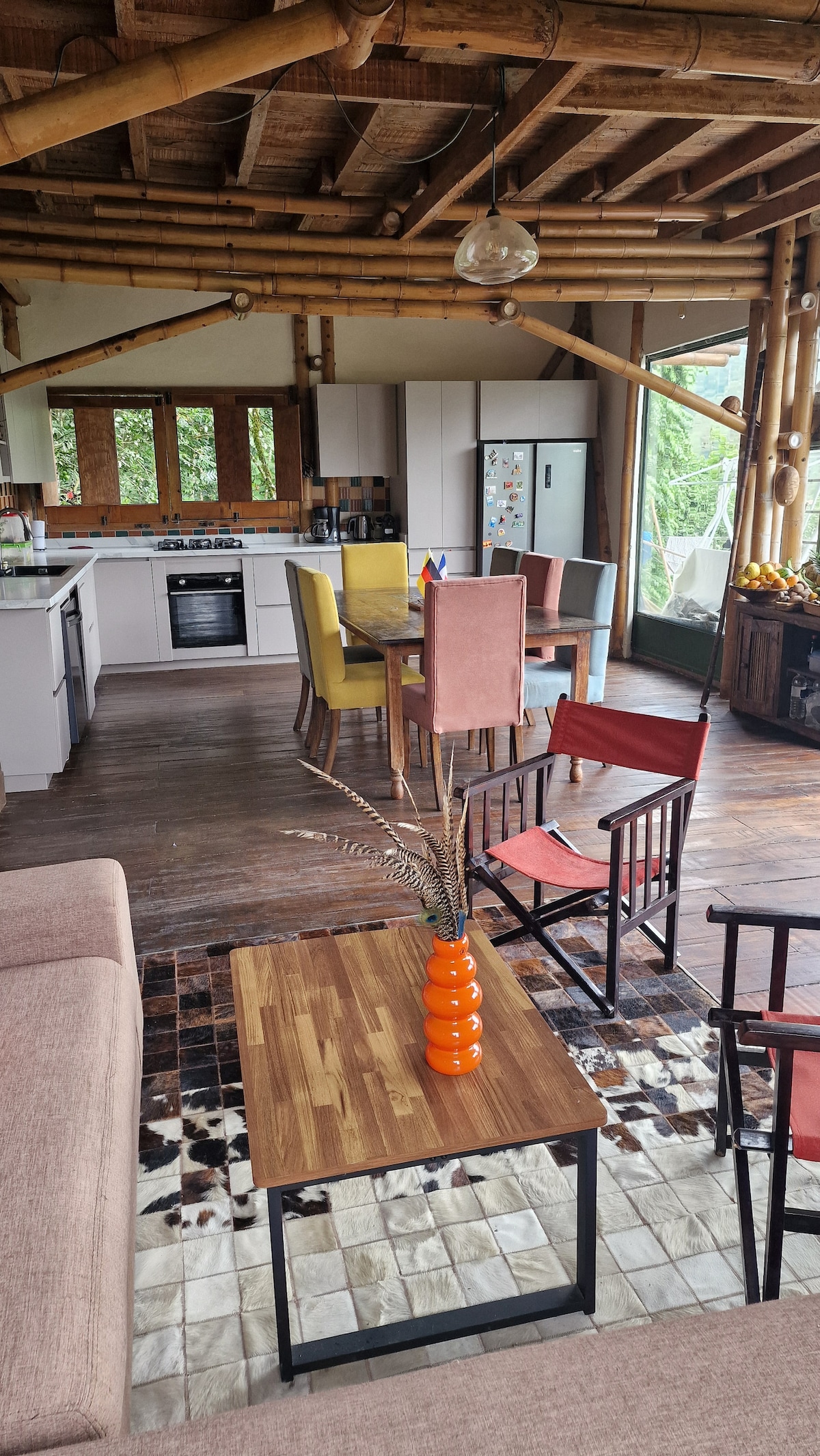 A cozy living space features a wooden table surrounded by colorful chairs. A sofa and minimalist coffee table sit nearby, while a modern kitchen area is visible in the background. Large windows allow natural light to fill the room, highlighting the bamboo ceiling beams.