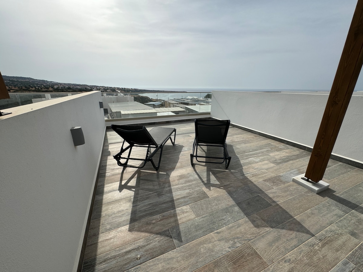 A private roof terrace features two black lounge chairs positioned to enjoy unobstructed views of the coastline. The flooring is composed of large tiles, and the surrounding areas are minimal, providing a serene space for relaxation.