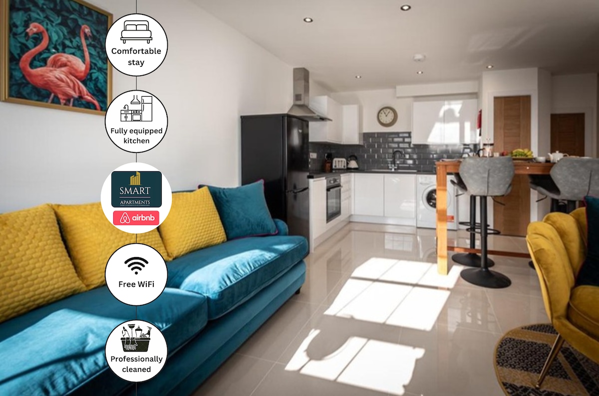 A modern living area features a comfortable blue sofa with yellow accent cushions. Natural light illuminates the space, highlighting a fully equipped kitchen in the background, which showcases sleek cabinetry and appliances. A dining area with high stools complements the overall contemporary design.