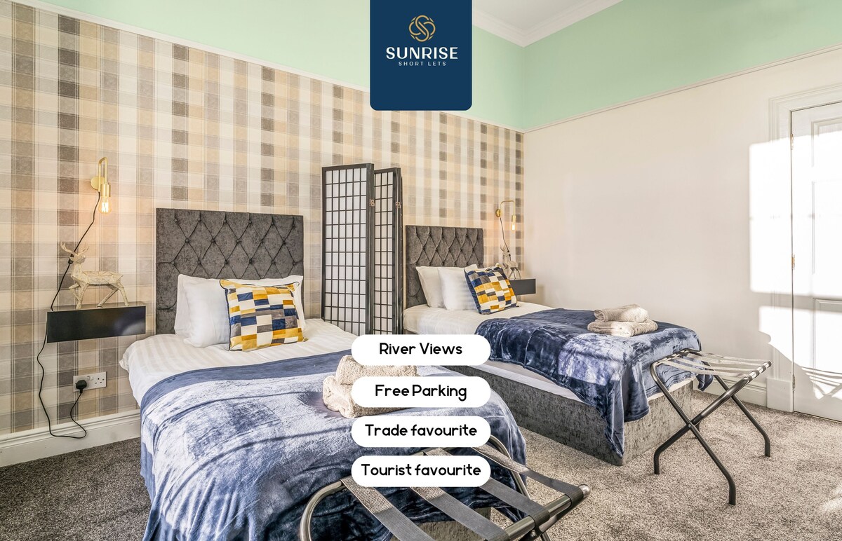 A well-lit guest room features two single beds with upholstered headboards, complemented by navy and white bed linens. A decorative folding screen adds privacy, while a patterned accent wall enhances the room's charm. Towels are neatly arranged on the beds, with a small side table positioned nearby.