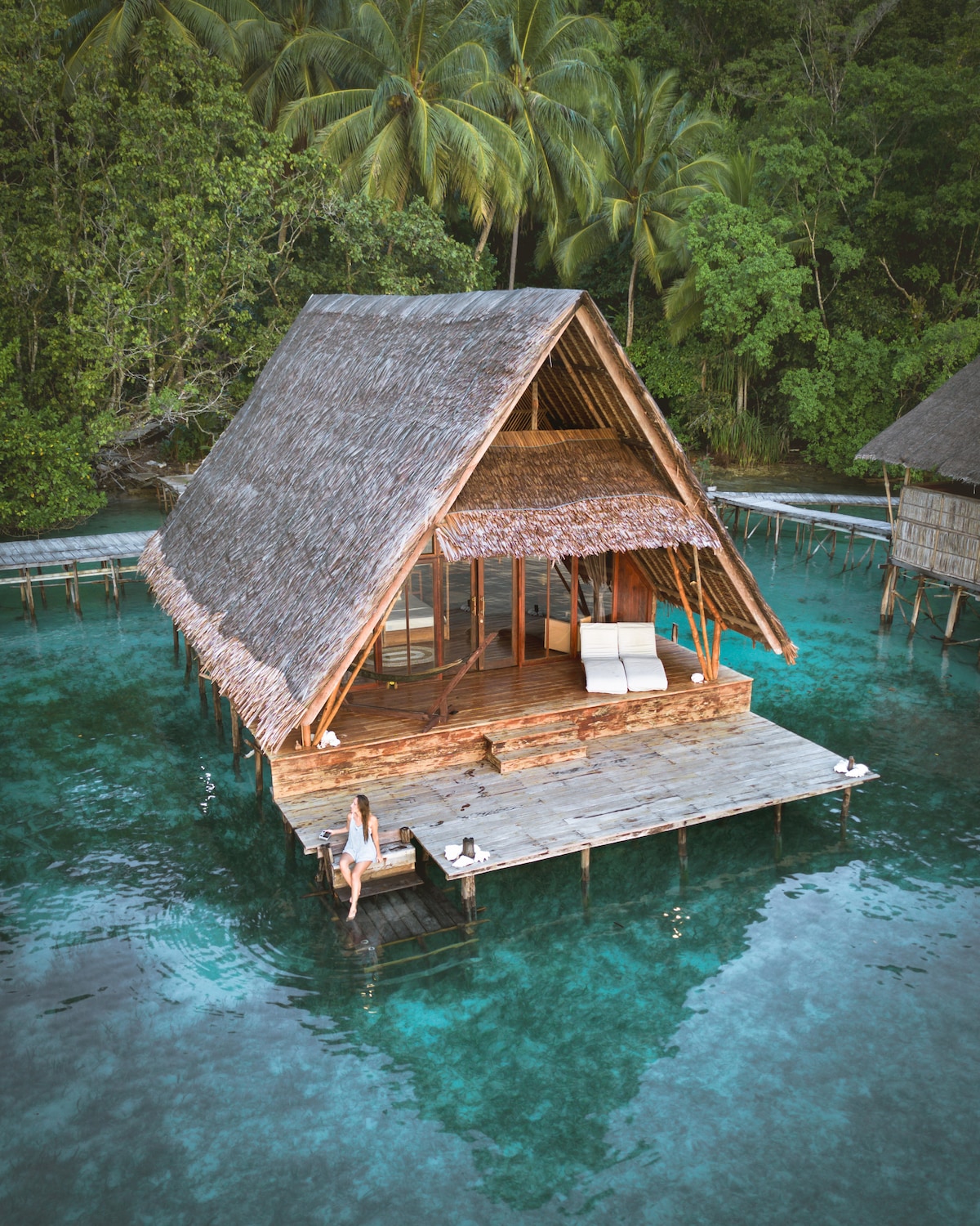 An overwater bungalow is set among calm, turquoise waters, framed by lush greenery. The structure features a traditional wooden roof and a spacious deck with sunbeds. A person can be seen relaxing on the edge, enjoying the serene surroundings.