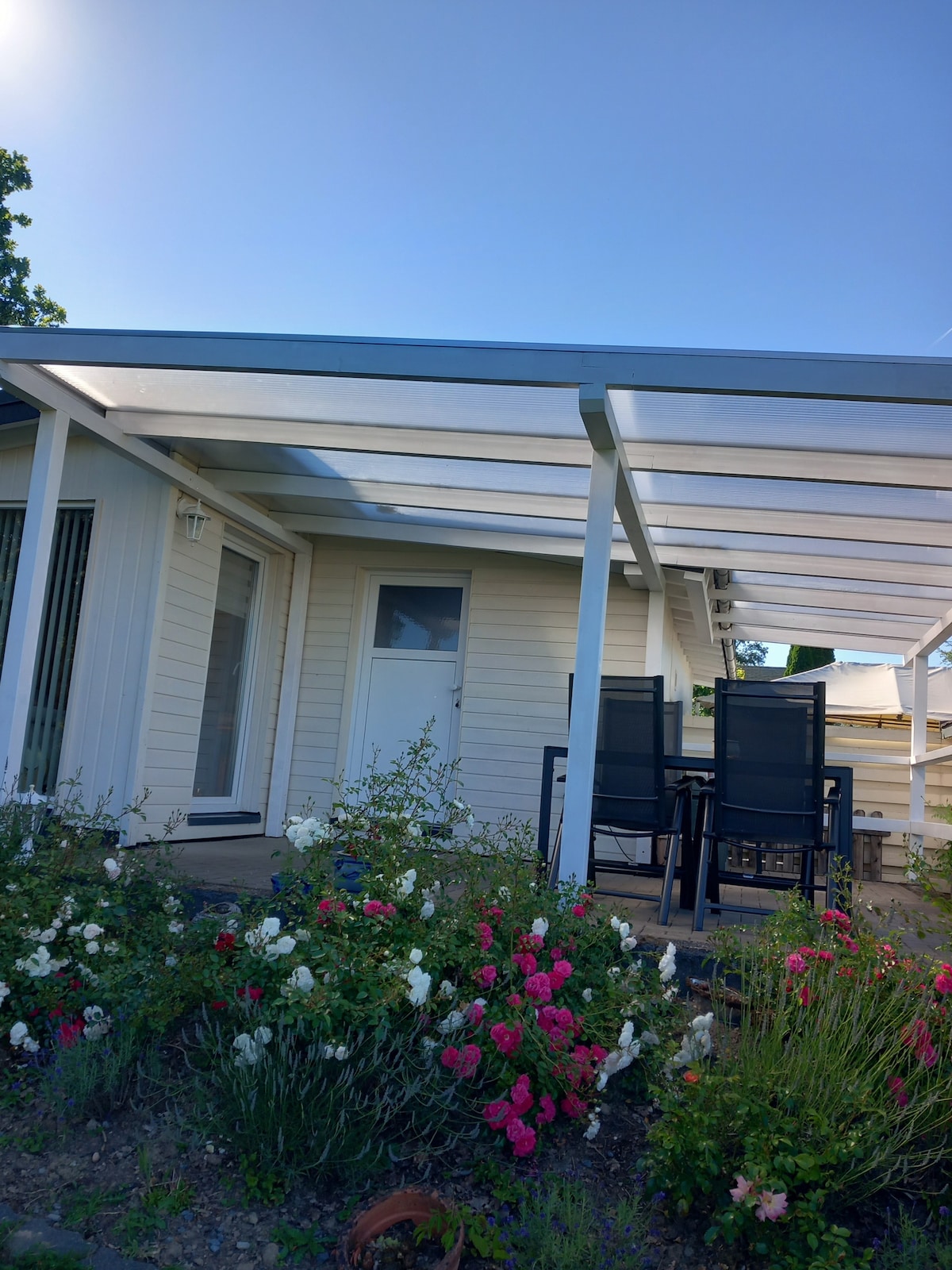 An exterior view of a small wooden house is shown, with a covered patio featuring a dining table and black chairs. Lush flower beds with pink and white blossoms frame the entrance, providing a colorful contrast to the light-colored façade.