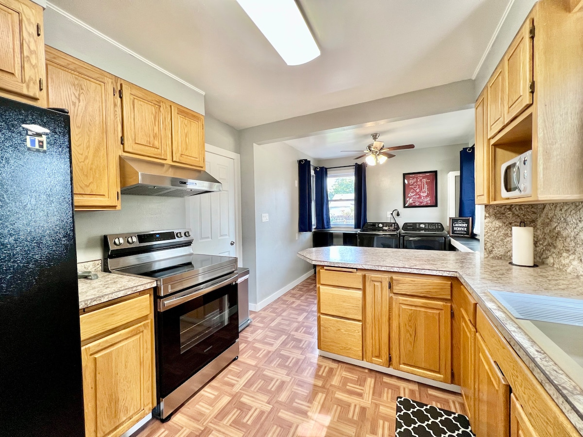 The kitchen features wooden cabinetry and a black stove with an oven. A spacious countertop allows for meal preparation, with various appliances visible. Natural light enters from a nearby window, and the floor is finished with patterned tiles. A laundry area is also present in the background.