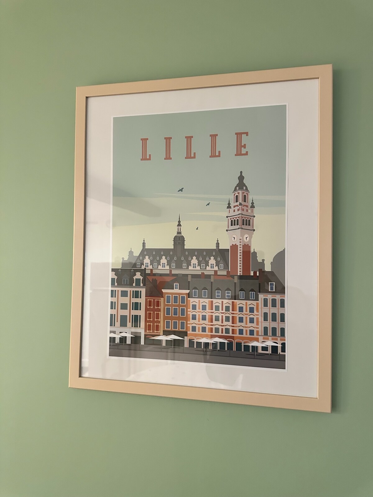 A framed artwork depicting a stylized view of Lille, featuring colorful buildings with intricate architectural details. The clock tower stands prominently, complemented by a clear sky with birds in flight. This piece adds a vibrant touch to the green wall.