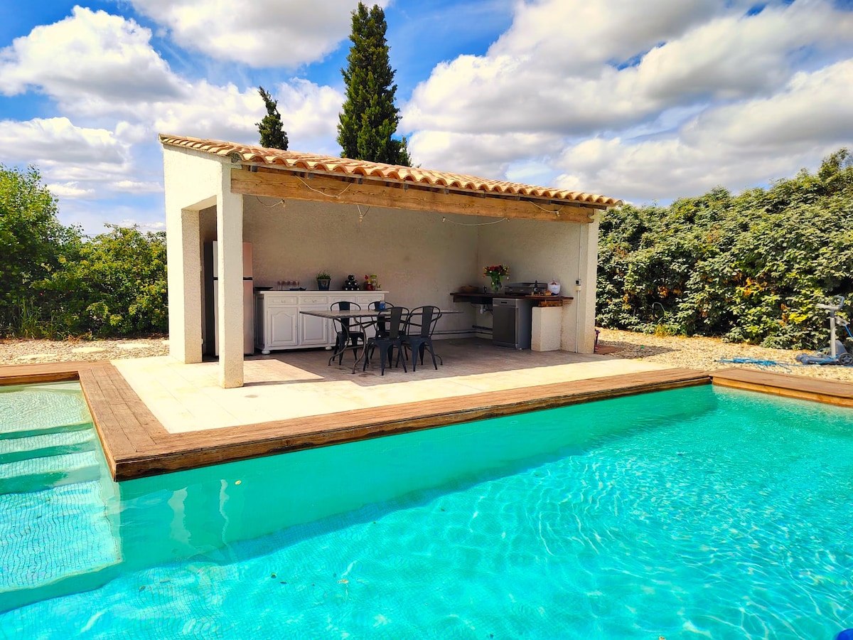 A shaded outdoor kitchen area is presented adjacent to a private pool, featuring a wooden deck. Several black chairs surround a table, and a barbecue grill is visible in the background. Lush greenery borders the space, enhancing the tranquil setting.