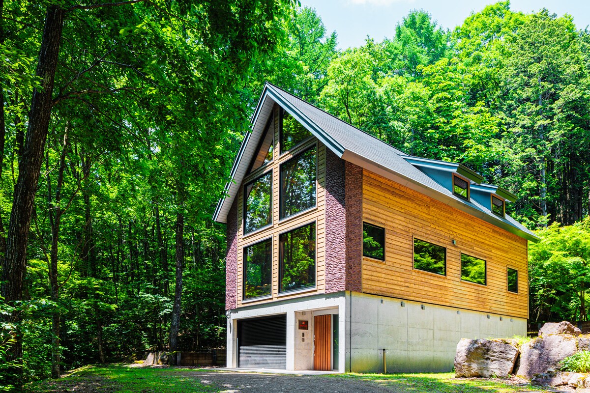 The cabin features a modern design with a combination of wooden and stone elements. Large windows provide views of the surrounding forest, allowing natural light to fill the space. The structure is nestled among lush greenery, creating an inviting and serene atmosphere.