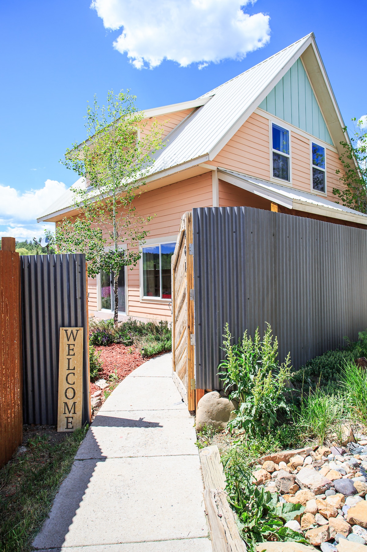 A welcoming pathway leads to the entrance of a charming home, flanked by landscaped greenery and rocks. A sign reading 'WELCOME' invites guests, while the house showcases a mix of pastel colors and architectural features under a bright blue sky.