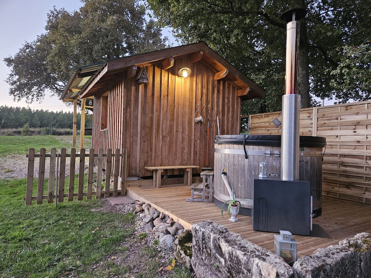 A wooden cabin is shown with a warm light illuminating its exterior. A wooden deck extends in front, and a hot tub is positioned beside the cabin. A rustic wooden fence encloses the area, with a backdrop of grassy open space and trees.
