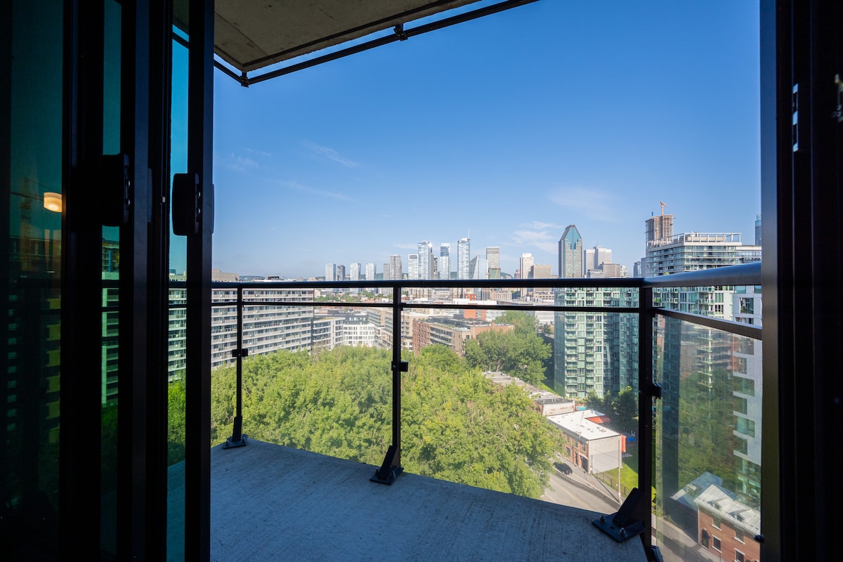 A private balcony is featured with a clear view of the city skyline, framed by glass doors. The surrounding area includes green trees, enhancing the urban atmosphere. The balcony's railing provides safety while allowing for an open vista of the horizon.