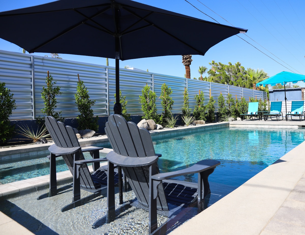 A serene saltwater pool is complemented by in-pool sun loungers, offering relaxation under a large shade umbrella. Lush greenery lines the poolside, enhancing the tranquil environment. The clean blue water reflects a clear sky, creating an inviting atmosphere for lounging.