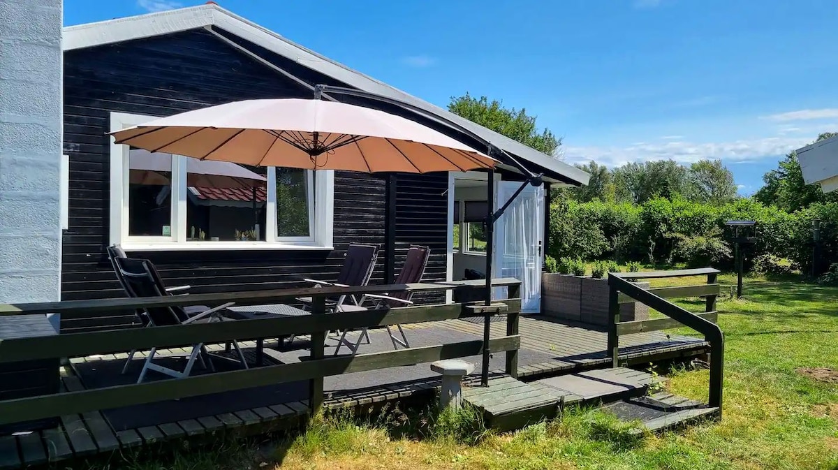 A charming cottage exterior features a large patio with a shaded umbrella and outdoor seating. The black wooden structure is complemented by large windows, offering views of the surrounding garden. Lush greenery borders the area, enhancing the tranquil setting.