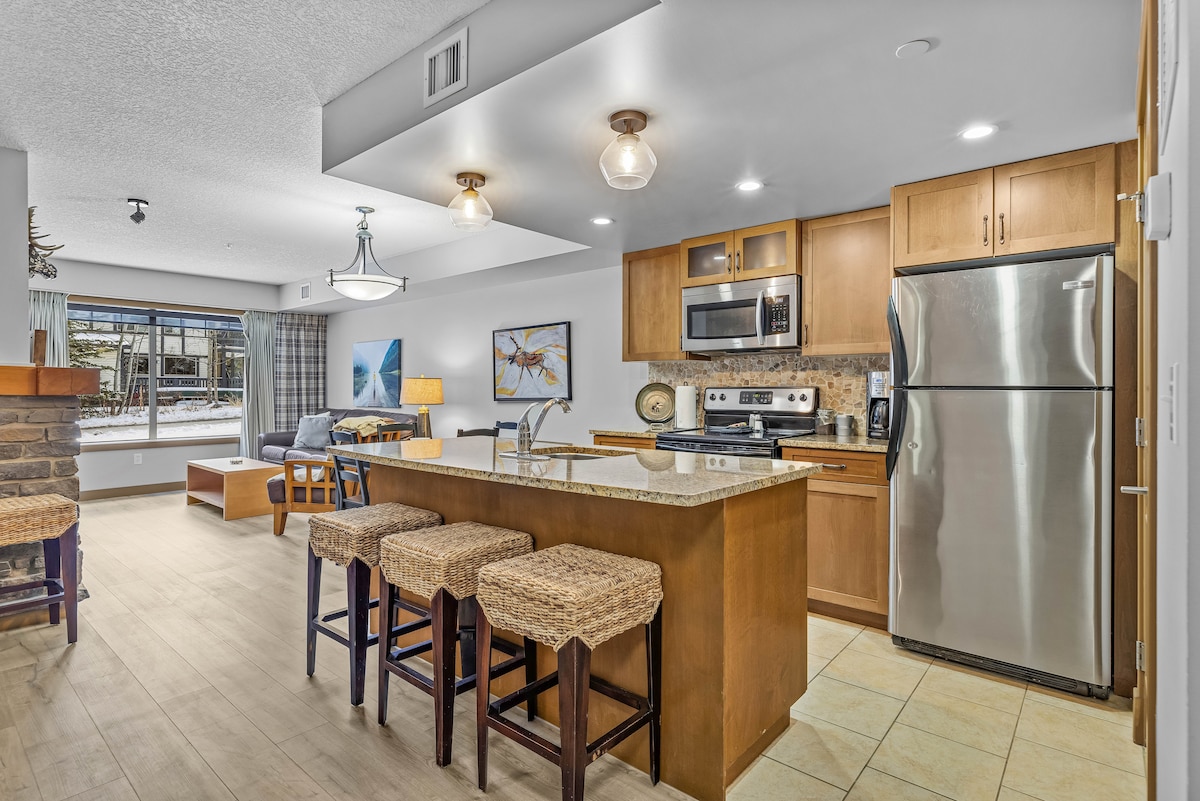 A modern kitchen area is visible, featuring stainless steel appliances and granite countertops. Bar stools are arranged at the kitchen island. Natural light filters in through large windows, illuminating the open living space. A cozy seating area with a sofa is positioned nearby.