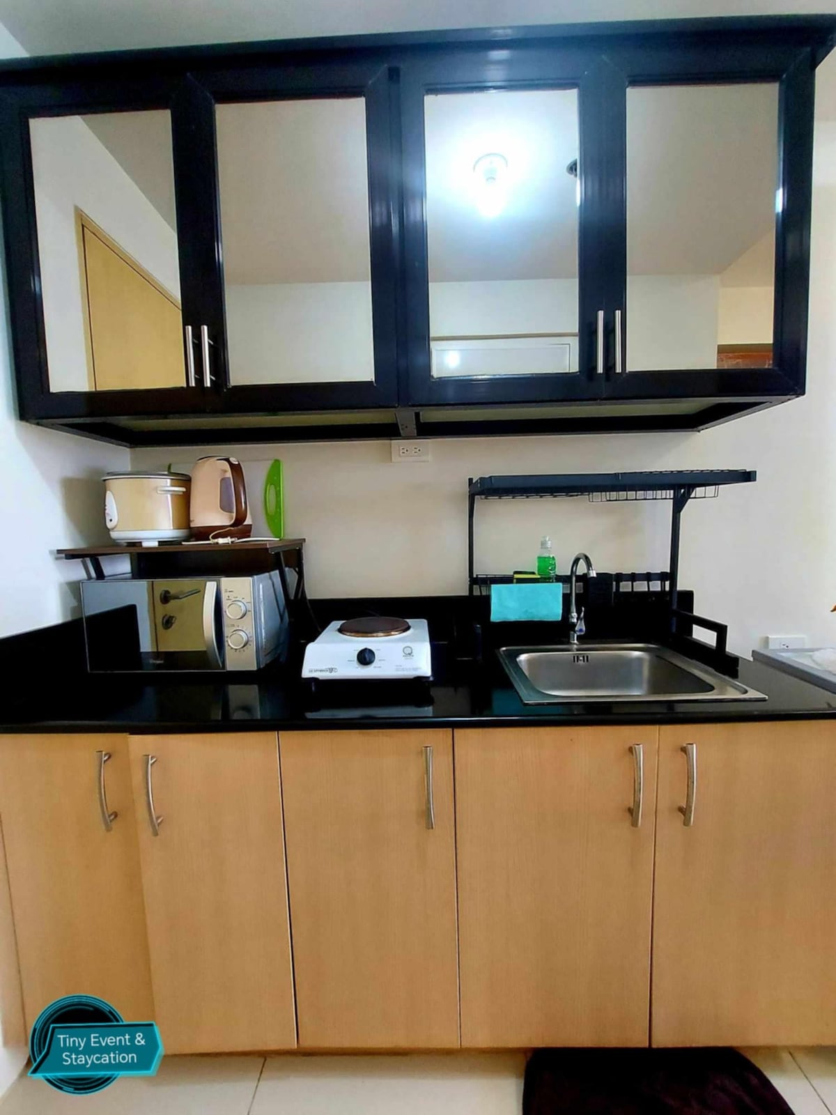 A compact kitchenette is featured, with wooden cabinets above and below. A black countertop hosts a sink, a stovetop, and a microwave, alongside a kettle. The space is well-organized, providing essential kitchen amenities for cooking.