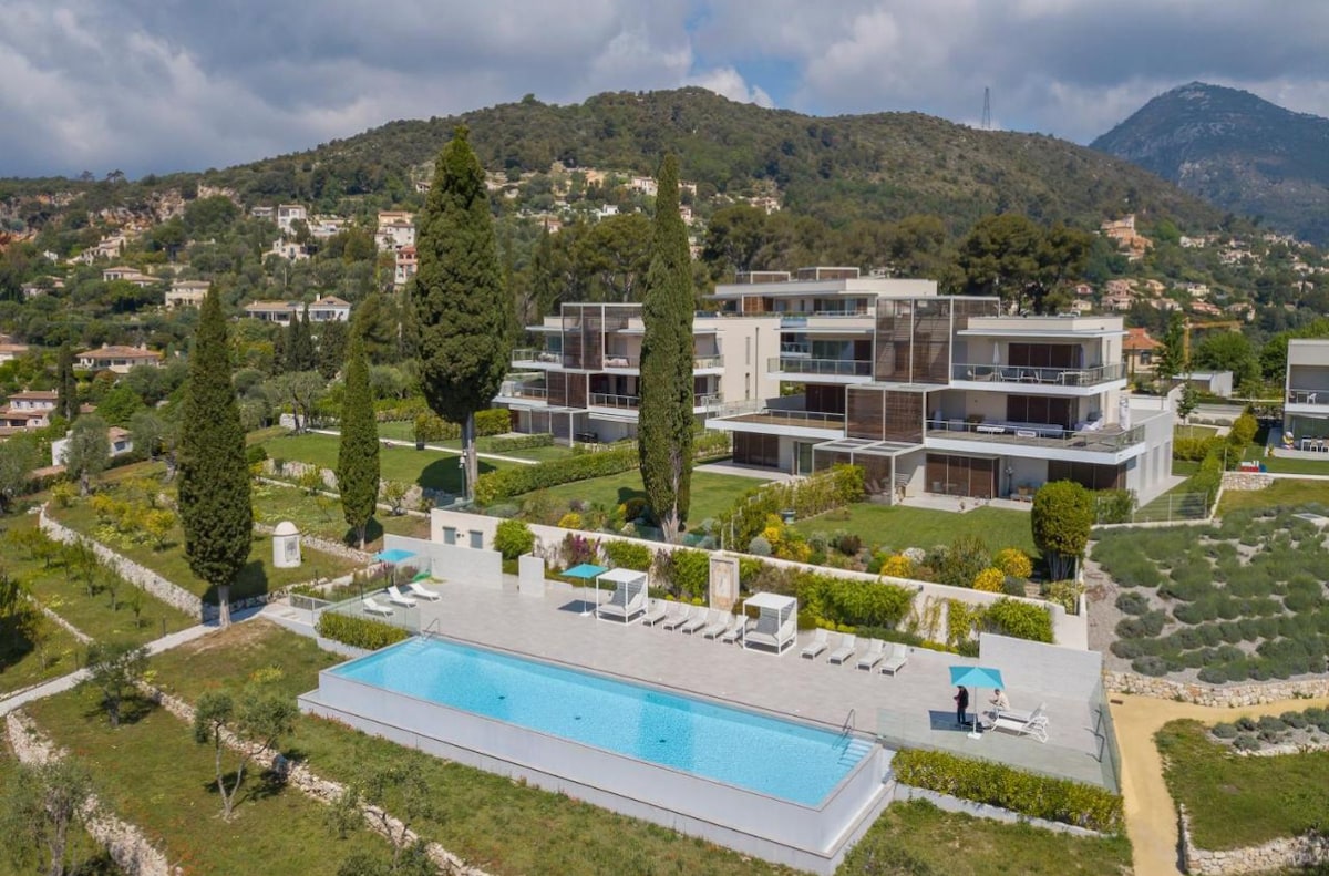 A modern residential complex is set amidst lush greenery, featuring multiple levels of outdoor terraces and large glass windows. An infinity pool with lounge chairs is positioned in the foreground, surrounded by manicured gardens. The backdrop includes rolling hills under a partly cloudy sky.