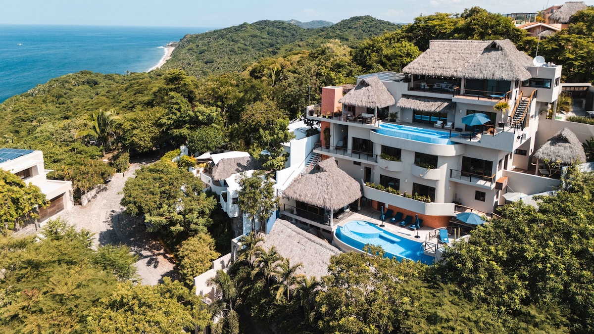 An aerial view of Casa El Mirador showcases its multi-level structure nestled within lush greenery. The property features several terraces, an inviting pool area, and the distant ocean, creating a serene atmosphere that blends seamlessly with the natural landscape.