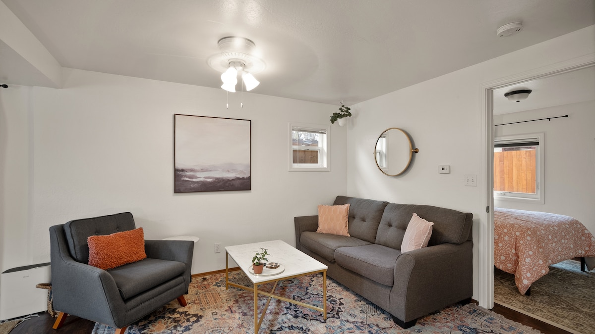 A cozy living area features a gray sofa with accent pillows and a matching armchair. A small coffee table sits in the center, adorned with greenery. Light walls are complemented by a round mirror and an abstract wall art piece, with a glimpse of a bedroom visible through an open door.