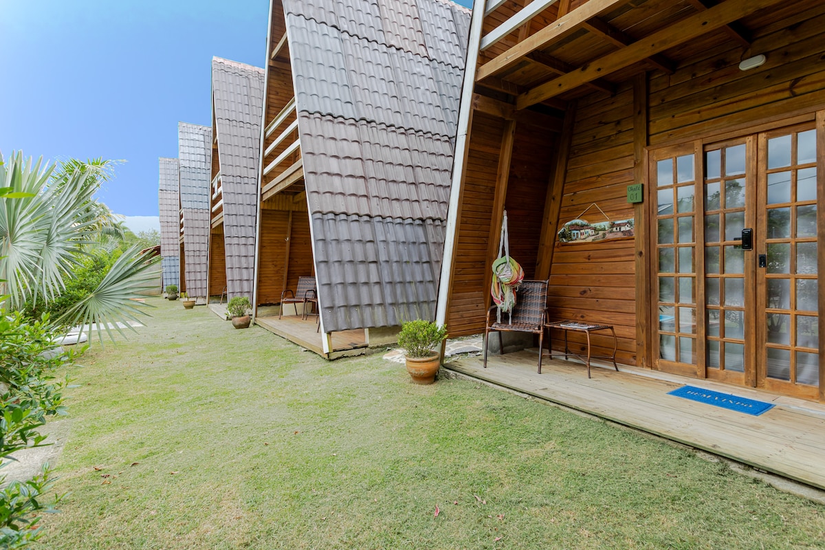 A row of wooden chalets with sloped roofs is set within a grassy area. Each chalet features a small porch with two chairs, potted plants beside the entrance, and a clear sky in the background, suggesting a serene outdoor environment.