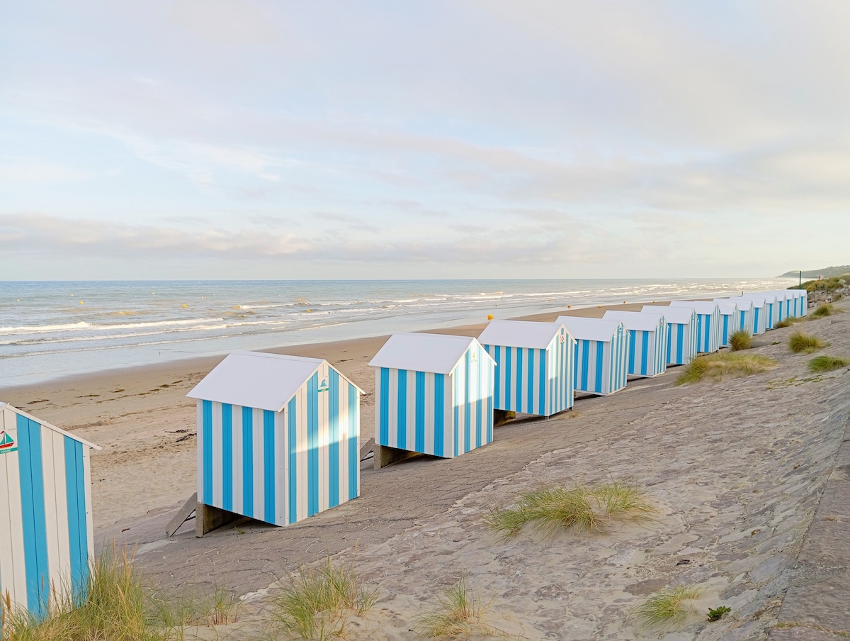 A row of white and blue striped beach huts is situated along a sandy beach. The gentle waves of the ocean can be seen in the background, while sparse grass and sandy areas surround the huts. A calm sky complements the beach setting.