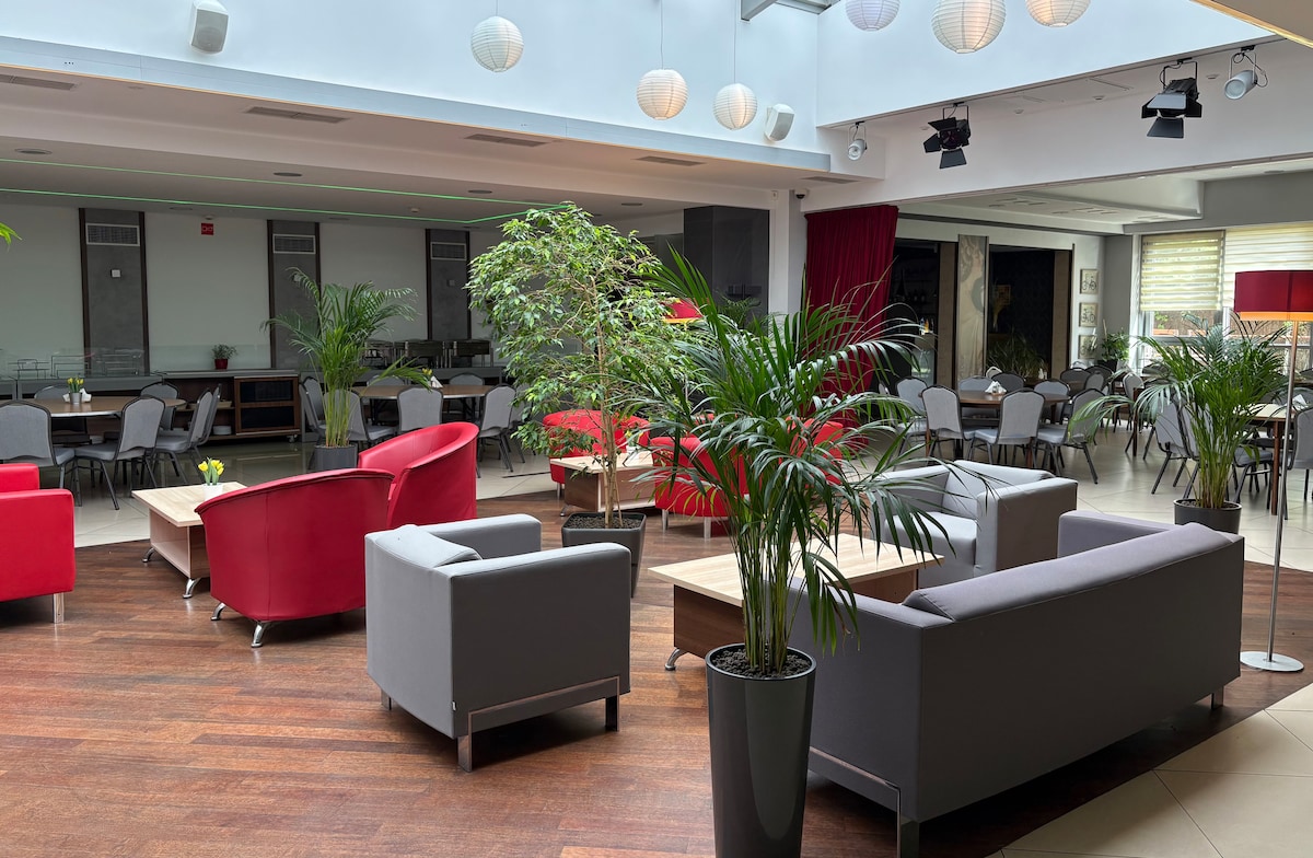 A spacious common area is characterized by modern furniture arrangements. Red and gray sofas are complemented by wooden coffee tables. Potted plants are placed throughout, adding a touch of greenery. Soft lighting from overhead fixtures enhances the inviting ambiance.