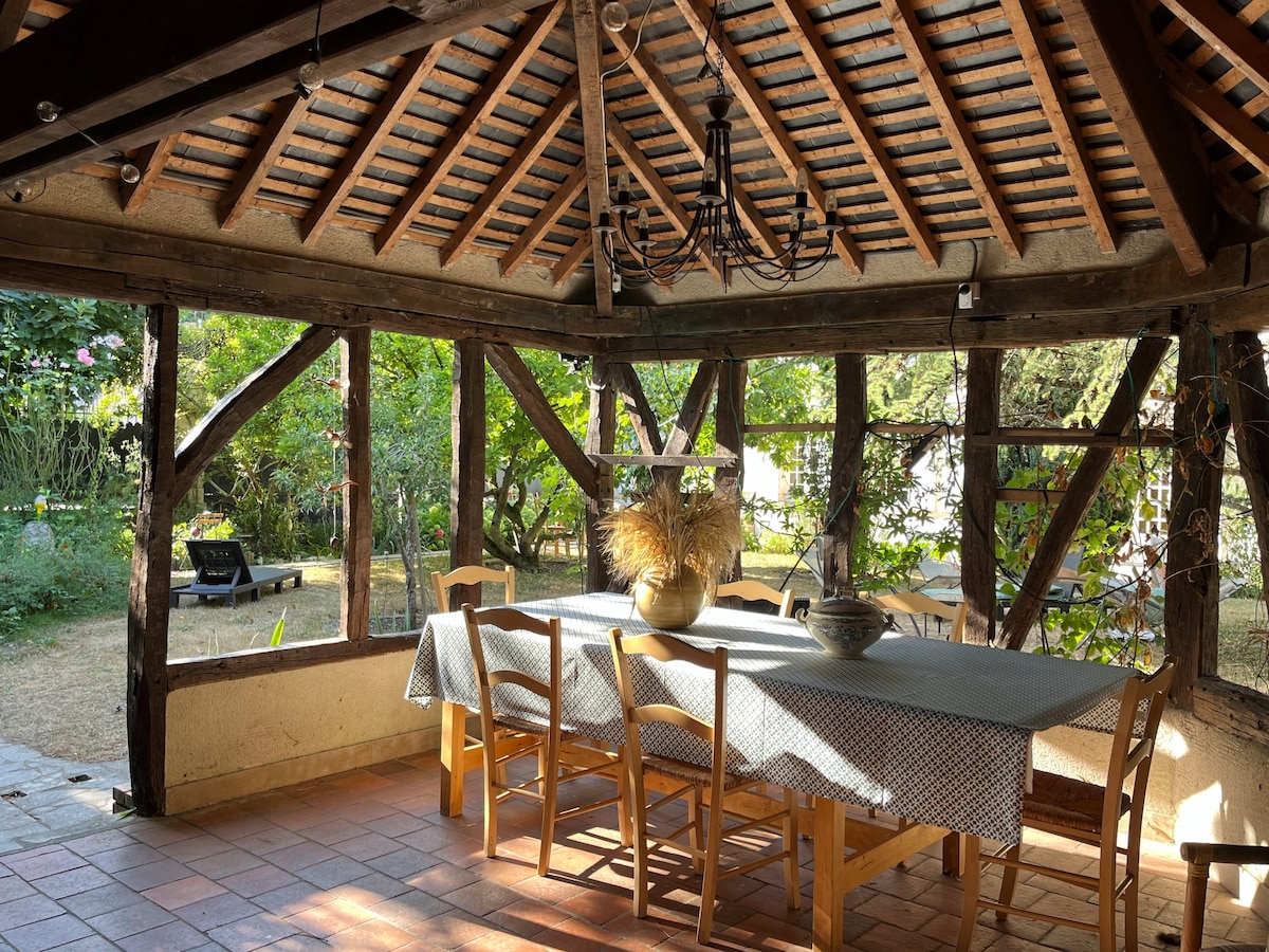 An outdoor dining area is set under a vaulted wooden ceiling with exposed beams. A large table is surrounded by several wooden chairs, and natural light filters through the open windows, illuminating the space. The surrounding garden is visible through the glass panels.