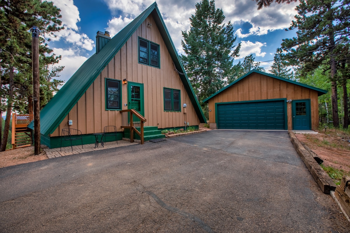 An A-frame structure is surrounded by tall trees and open sky, featuring a multi-colored exterior with green and brown tones. A paved driveway leads up to the house, with steps ascending to the front porch and an attached garage visible to the side.