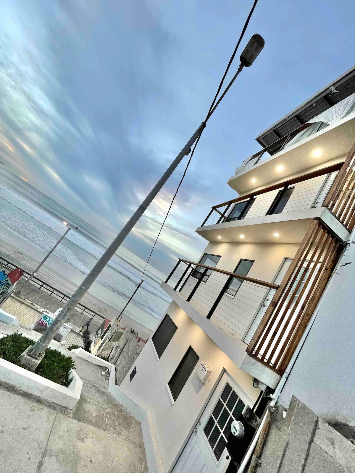 The exterior of a three-story apartment building is showcased, with sleek white walls and wooden accents. A staircase leads down towards the beach, while the ocean waves can be seen in the background, under a softly lit sky transitioning into evening.